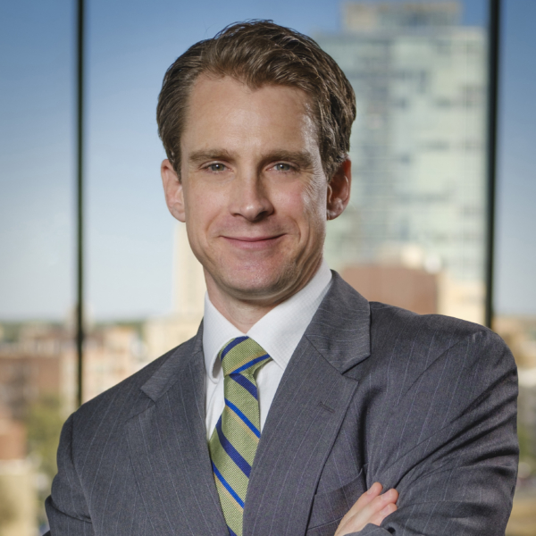 Andrew Holton, the new COO of Research Triangle Foundation, standing confidently with arms crossed, wearing a gray pinstripe suit, a white shirt, and a green striped tie. He is smiling with a city skyline blurred in the background, viewed through a large window.