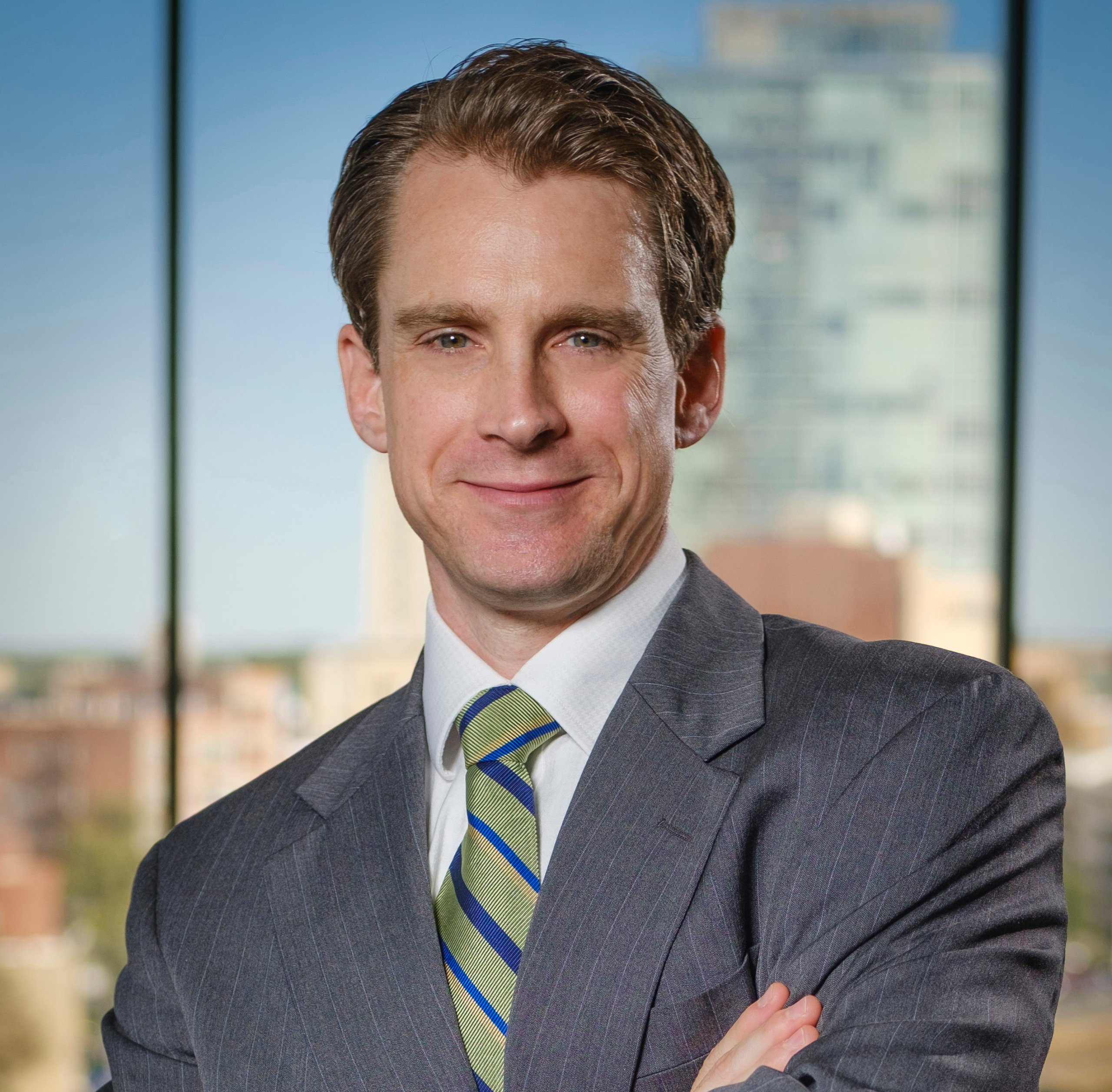 Andrew Holton, the new COO of Research Triangle Foundation, standing confidently with arms crossed, wearing a gray pinstripe suit, a white shirt, and a green striped tie. He is smiling with a city skyline blurred in the background, viewed through a large window.