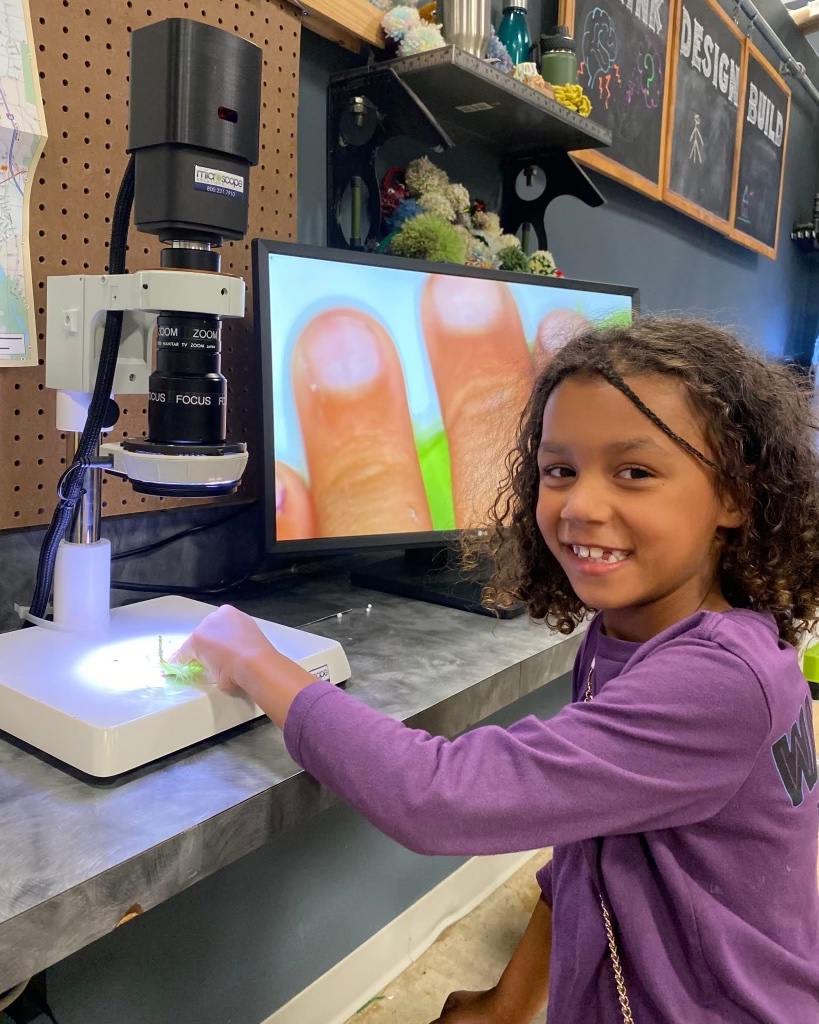 A young girl with curly hair smiles while examining a small object under a digital microscope. The microscope's screen shows an enlarged view of the object and part of her hand.