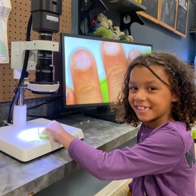 A young girl with curly hair smiles while examining a small object under a digital microscope. The microscope's screen shows an enlarged view of the object and part of her hand.